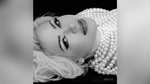 Anelise poses with a close-up headshot, wearing heavy pearl necklaces and large pearl earrings against a black background.