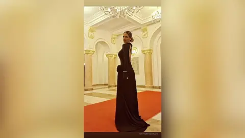 TiffanyAddams poses in a long, black backless gown while standing on a red carpet in a bright, white room with gold columns.