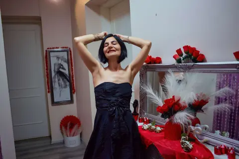 Cheerful woman in a black strapless evening dress posing in a room with red floral decor.