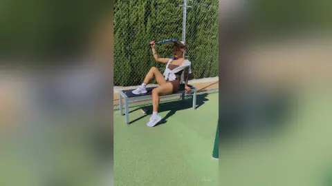 Young woman in casual summer outfit sitting on a bench at an outdoor tennis court on a sunny day.