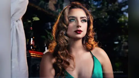 Glamorous woman with wavy brown hair in a green dress posing in a moody, dimly lit setting.