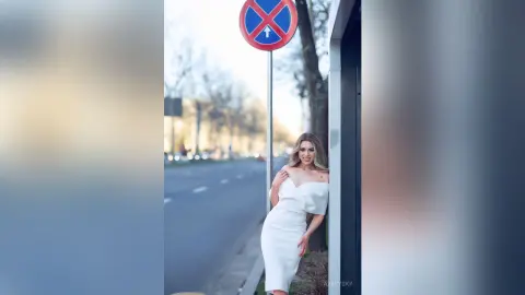 Smiling woman in an elegant white cocktail dress leaning against a wall on a sunny city street.