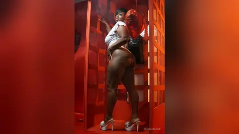 Curvy woman in white top and heels posing in a red room with a heart balloon for a romantic mood.