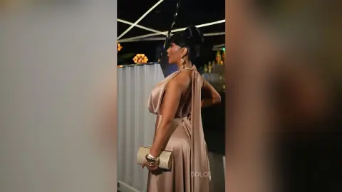Dolce poses in a tan sleeveless dress with a long drape, wearing gold earrings and holding a gold clutch.