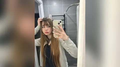 Young woman with long brown hair taking a mirror selfie in a modern bathroom wearing a beige jacket.