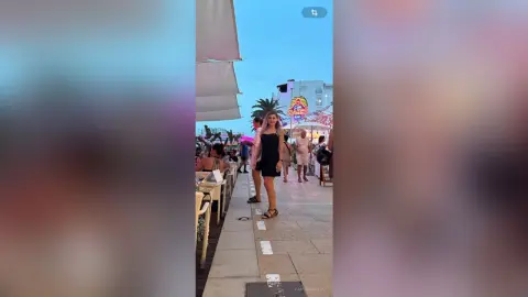 Young woman in a black summer dress standing at an outdoor seaside cafe during a breezy twilight evening.