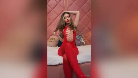 Glamorous woman in a red lace bodysuit posing on a pink bed in a luxurious bedroom setting.