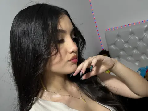 Young woman with long dark hair wearing a white top in a cozy bedroom setting.