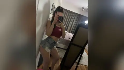 Young woman in a red crop top and denim shorts taking a mirror selfie in a modern bedroom.