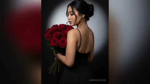 LaurenKaterine poses with a large bouquet of red roses, wearing a black spaghetti-strap dress with her hair in a bun against a dark background.