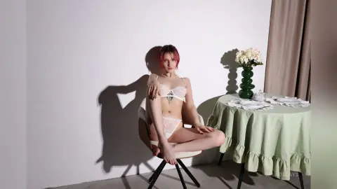 Young woman with pink hair in white lace lingerie sitting on a chair in a bright minimalist room.
