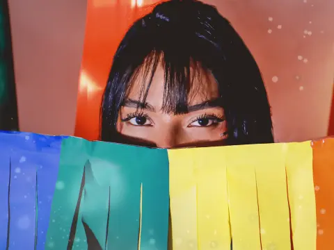 EmmaEbonyX19 peeks over a colorful rainbow paper banner, showing her dark hair with bangs and her eyes looking directly at the camera.