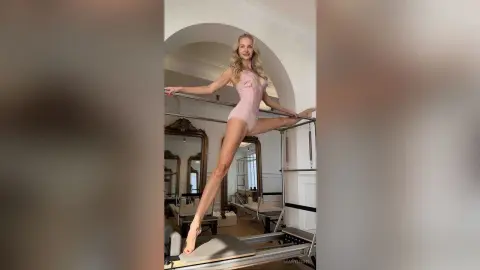 Blonde ballerina in a pink leotard performing a graceful leg stretch in a bright dance studio.