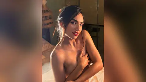 AlessiaMoon poses naked in a bedroom, using one hand to cover her breast while looking at the camera with long dark hair.