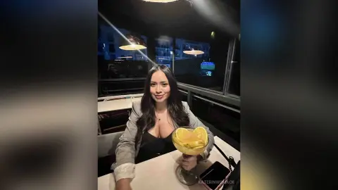 Smiling woman in a black top and grey blazer enjoying a cocktail at a dimly lit restaurant at night.