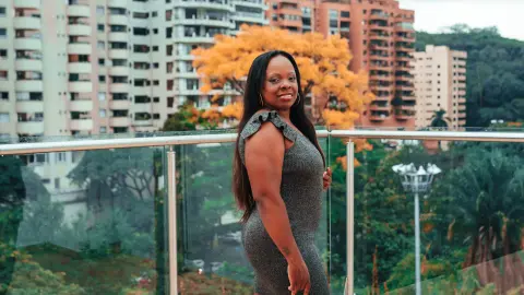 Smiling woman in a grey sleeveless dress standing on a balcony overlooking city buildings and lush greenery.
