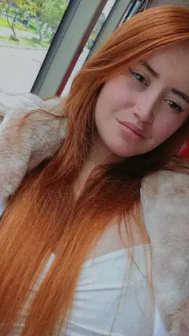 Redheaded woman in a white top and fuzzy beige coat smiling softly by a window.