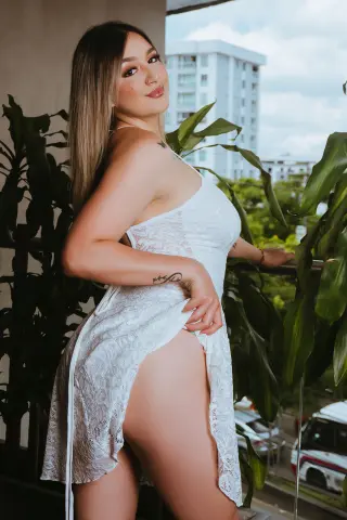 AnnieLeonii poses on a balcony wearing a white lace slip, lifting the hem to show her thigh and hip against a background of green leaves and buildings.