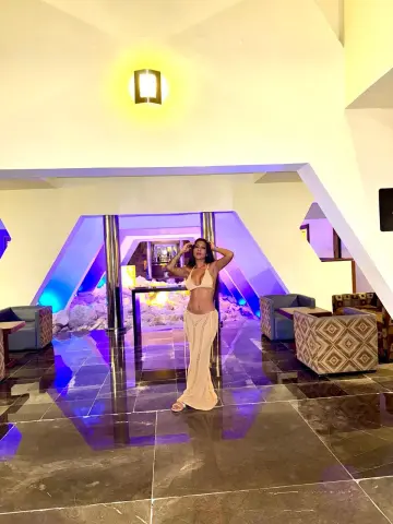 Stylish woman in a white bikini and maxi skirt posing in a luxury modern hotel lobby with blue ambient lighting.