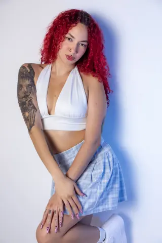 Young woman with curly red hair in a white halter top and blue plaid skirt posing against a white background.