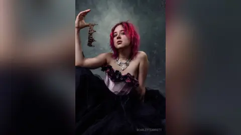 ScarlettGarrett poses in a black and pink corset with pink hair, holding a gold decorative piece against a dark grey background.