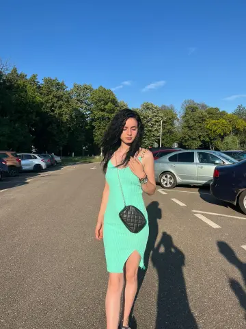 Young woman in a mint green summer dress and black crossbody bag standing in a sunny outdoor parking lot.