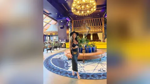 ArianaKroft stands in a luxury lounge wearing a black lace bra and matching black skirt with a black wide-brimmed hat.