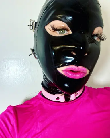 Woman in a black shiny latex mask and pink collar wearing a bright pink top against a white background.