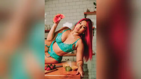 IsabellaEstrada poses in a bright kitchen wearing a blue lace lingerie set, holding a slice of watermelon near her head with her red hair down.
