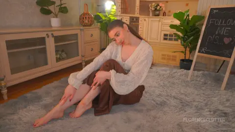 Young woman in a white bohemian blouse and brown trousers sitting on a shag rug in a cozy, sunlit living room.