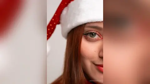 ArinnaGreen wears a red and white Santa hat and red eye makeup, looking toward the camera with long red hair.