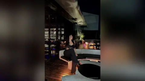 Stylish woman in a black crop top and trousers sitting on a lounge sofa at a moody rooftop bar at night.