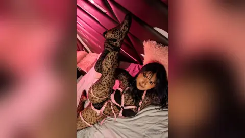 Woman in black lace bodysuit and patterned tights relaxing on a bed in a pink moody bedroom setting.