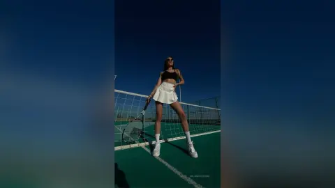 MilanaVolkova stands on a tennis court wearing a black bra and a short white skirt, holding a tennis racket against a clear blue sky.
