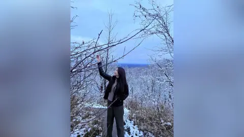 Woman in a black leather jacket standing in a snowy winter forest reaching toward bare tree branches.