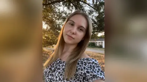 Smiling blonde woman in a black and white floral blouse relaxing outdoors in a sunny park setting.