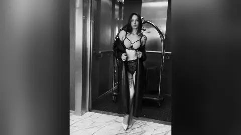 Elegant woman in black lace lingerie and a long robe posing in a moody, black and white hotel hallway.