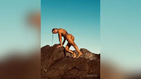 Fit woman in black bikini posing on rocky cliffs under a clear blue sky during a sunny summer day.