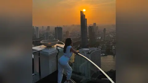 AnaMathers stands on a high balcony overlooking a city skyline at sunset, wearing a white sleeveless jumpsuit.