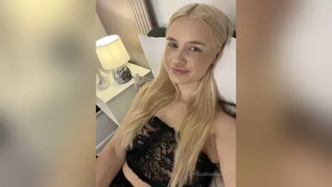 Smiling blonde woman in a black lace top relaxing on a white pillow in a cozy, dimly lit bedroom.