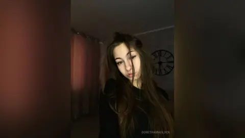 Young woman with long brown hair wearing a black top in a dimly lit room with a vintage wall clock.