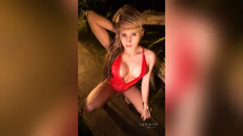 AndreaKalo wears a red lace bodysuit and a gold headpiece while posing outdoors in a dark, natural setting.
