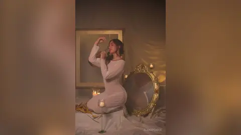 AlanaPearce poses in a white long-sleeve dress, sitting on white sheets next to a gold mirror and lit candles.