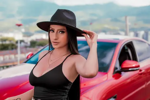 Stylish woman in a black tank top and wide-brimmed hat posing confidently next to a red car outdoors.