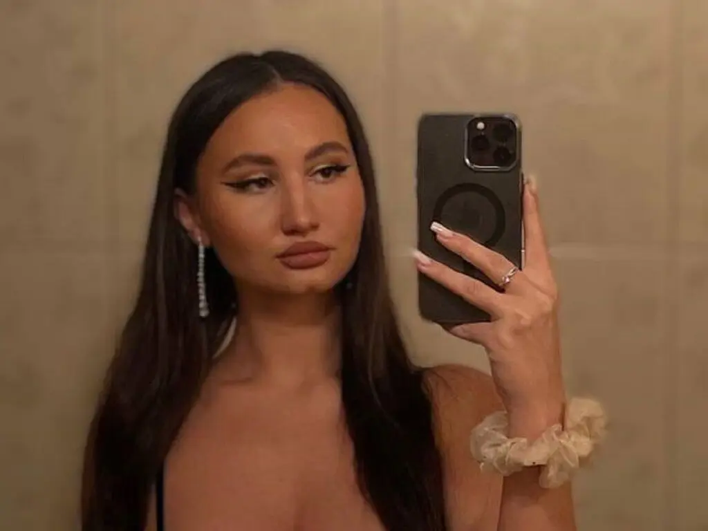 Young woman with long dark hair taking a moody mirror selfie in a neutral setting.