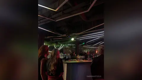 SabrinaSteels stands with her back to the camera in a dimly lit bar setting, showing her long blonde hair and dark top.