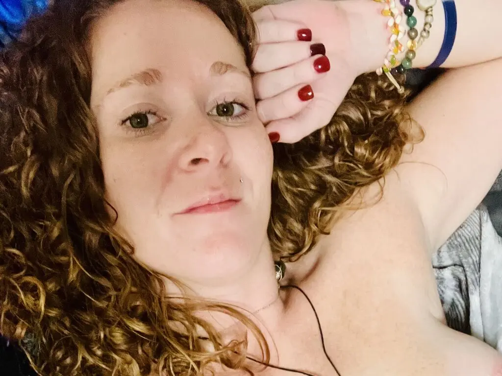 Smiling woman with curly brown hair and red nails relaxing in a cozy indoor setting.