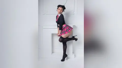 Young woman in a schoolgirl outfit with a pink plaid skirt and black tights posing in a bright white studio.