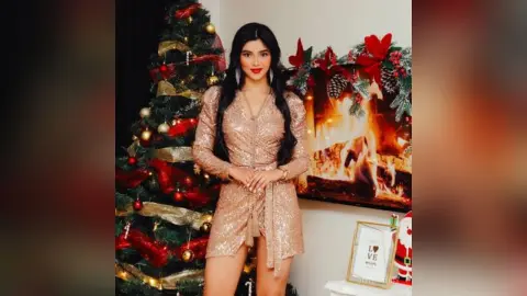 Young woman in a sparkling gold sequin dress posing by a decorated Christmas tree and cozy fireplace.