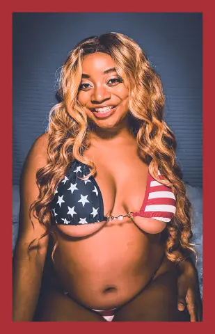 Smiling woman with long wavy blonde hair wearing a star and stripe bikini in a soft indoor setting.
